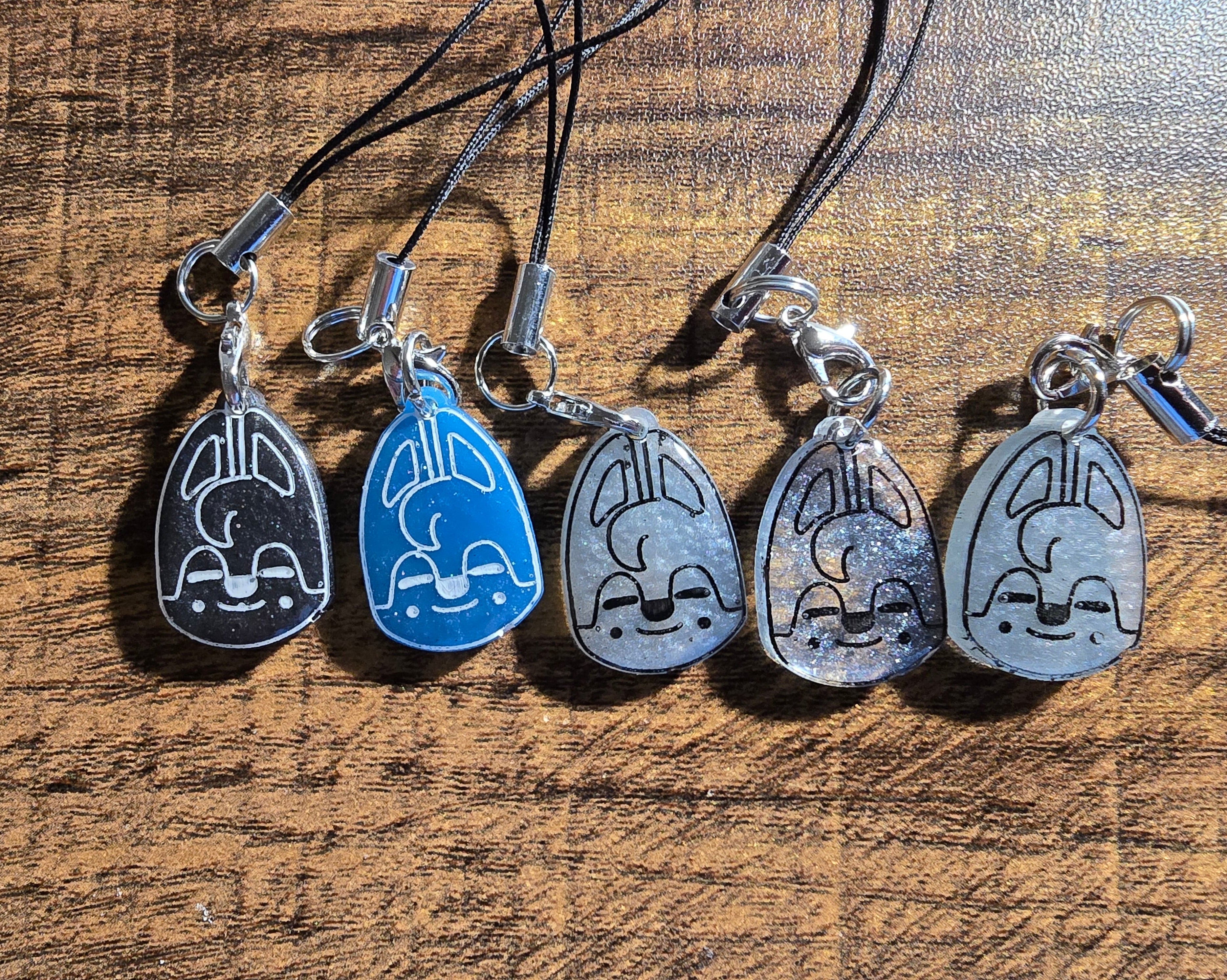 Five wolfchan shaped resin charms  on black cords against a wooden background. Charms are of varying blue to grey colors